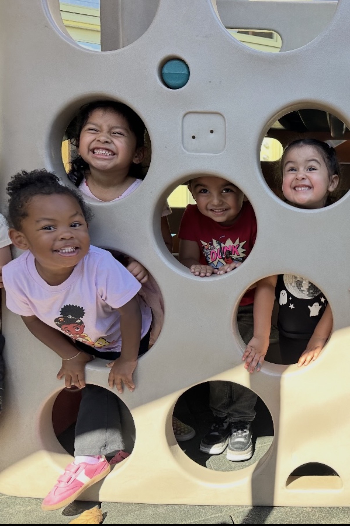 Four preschoolers smiling through the round windows of the play structure